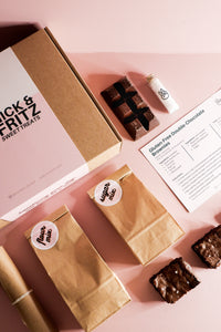 Vertical photo showing contents of Nick&Fritz Gluten-Free Brownie Baking Kit, incl. box, chocolate, sea salt, flour & sugar mixes, sample brownies, & recipe